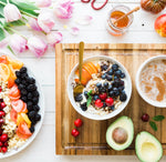 Tagliere con due ciotole, avocado, frutta, ciliegie, porridge, Piatto con mirtilli, ciliegie, pesca e porridge, mela, tazza con miele, fiori